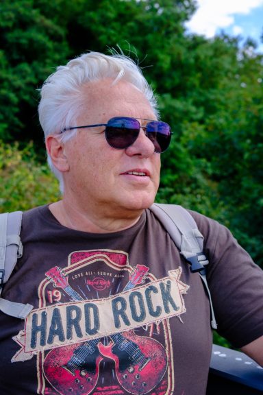 Mann mit grauen Haaren und Sonnenbrille, trägt ein T-Shirt mit "Hard Rock"-Aufdruck.