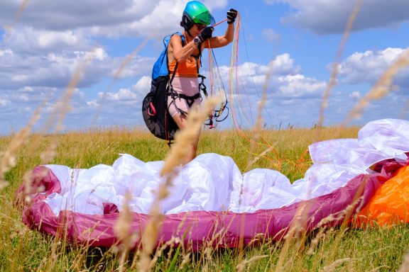 Person mit Helm bereitet einen Gleitschirm auf einer Wiese vor; Wolkenhimmel im Hintergrund.