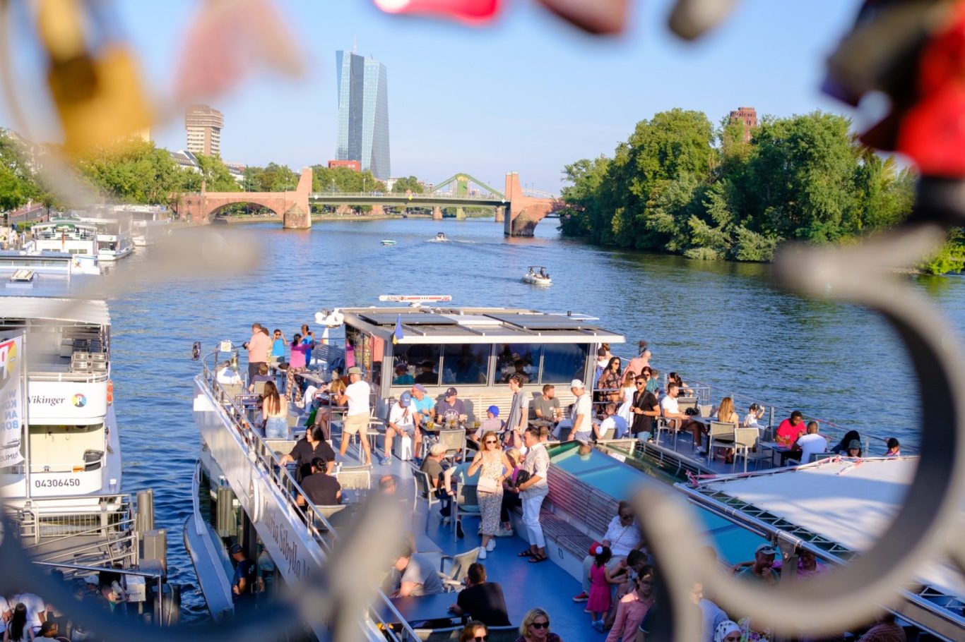 Eventfotografie Blick auf ein voll besetztes Boot auf einem Fluss mit Stadtlandschaft im Hintergrund.