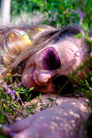 Frau mit Sonnenbrille liegt lächelnd im Gras zwischen bunten Blumen.