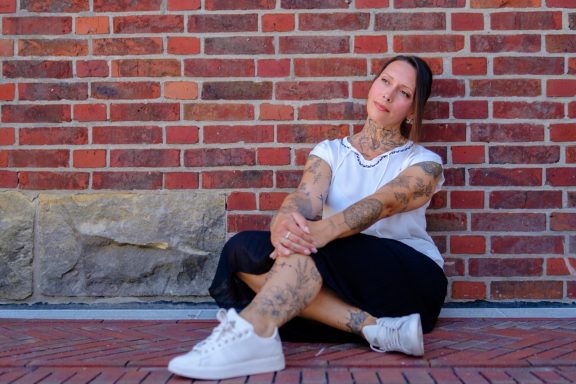 Frau mit Tattoos sitzt lässig vor einer Backsteinwand.