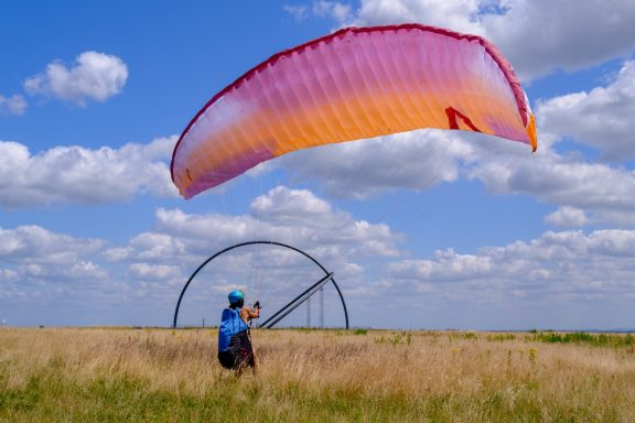 Ein Paraglider mit buntem Schirm steht in einer Wiese unter einem bewölkten Himmel.